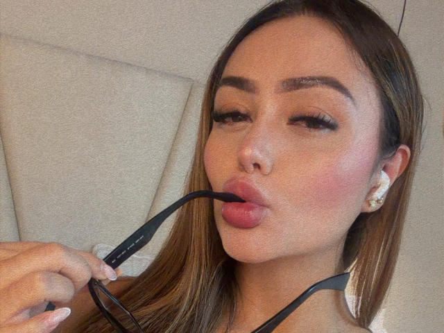 Close-up of MissAry with long brown hair, holding black straps near her lips, which are slightly puckered. She has full eyebrows, pink lips, and white earrings. Slight sexual tension.