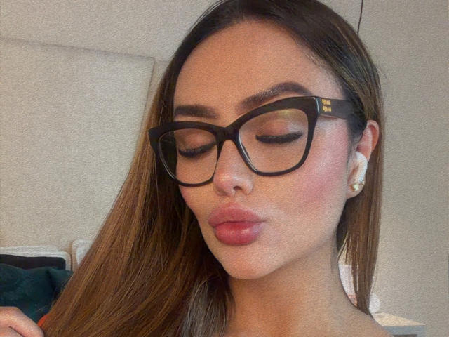 Photo en gros plan de MissAry, avec de longs cheveux bruns, des lunettes à monture noire et du rouge à lèvres rose. Elle fait la moue, les yeux fermés, et porte des boucles d'oreilles en diamants. Incroyablement sexy, avec des sourcils épais, une lumière douce et un éclat subtil..