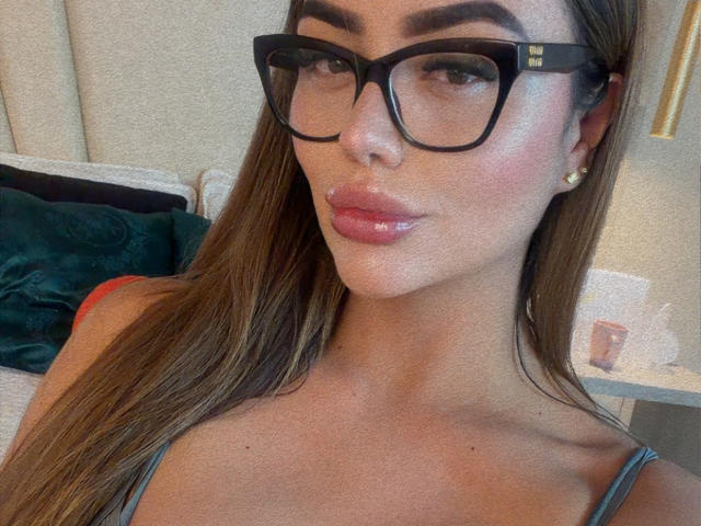 Close-up of MissAry with glossy pink lips, large black-framed glasses, and long brown hair. She has a sultry expression, wearing a gray tank top, and small gold earrings. Fucking sexy.