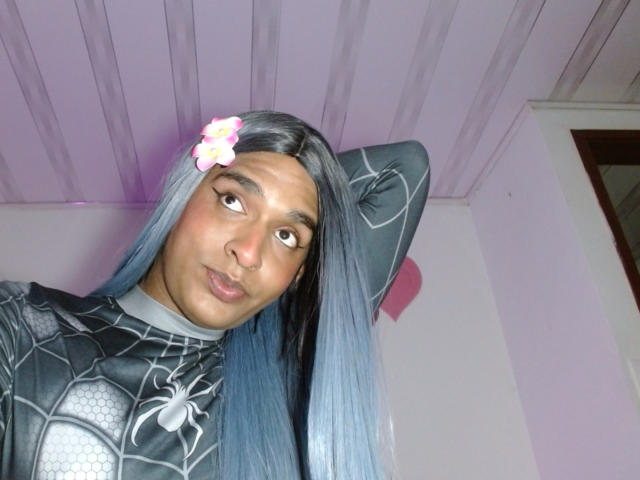 Fotografía de IdiiaLatina con cabello largo, lacio y azul oscuro, vestida con un traje negro de Spider-Man, con una mano detrás de la cabeza, de color blanco. Calcomanía de flor en la frente, ojos rosados, labios ligeramente fruncidos, techo morado arriba..