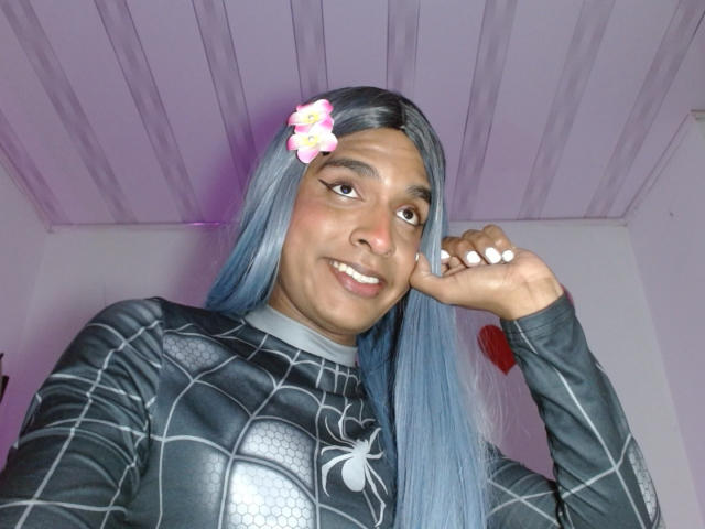 Fotografia de IdiiaLatina com cabelo azul comprido, vestindo uma camiseta preta do Homem-Aranha, sorrindo para a câmera, com a mão tocando [algo]. bochecha, prendedor de flor no cabelo, teto branco e claro acima. Muito fofo.