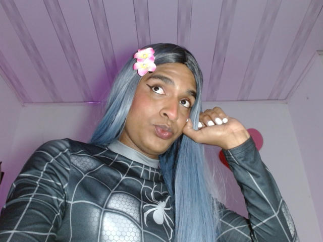 Photograph of IdiiaLatina with long blue hair, wearing a black checkered shirt with a white spider emblem. She's posing with hand on cheek, pouty lips, purple ceiling background. Fucking sexy.