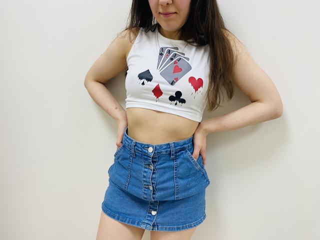 JoannaSia, avec de longs cheveux bruns, portant un crop top blanc avec un motif de cartes à jouer et des cœurs rouges, et une jupe en jean bleu, debout, les mains dans les poches, fond blanc, léger sourire..
