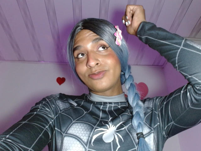 IdiiaLatina con una larga trenza azul, vistiendo un traje negro de Spider-Man, sonriendo, con la mano levantada, iluminación púrpura, símbolos de corazón. Traje ajustado que realza la forma del pene, emblema de araña blanca en el pecho, pose segura..
