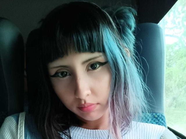 Close-up of KleoLennox, a woman with dark blue hair in two buns, heavy black eyeliner, and pale skin. She's looking directly at the camera with a neutral expression. Bright light from the right.