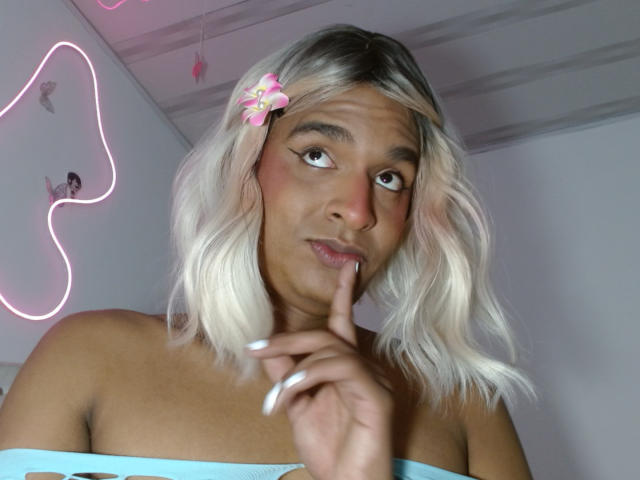 Close-up of IdiiaLatina with platinum blonde hair, pink flower clip, and blue strapless top. She's shushing with a finger to her lips, neon pink outline on the wall in the background.