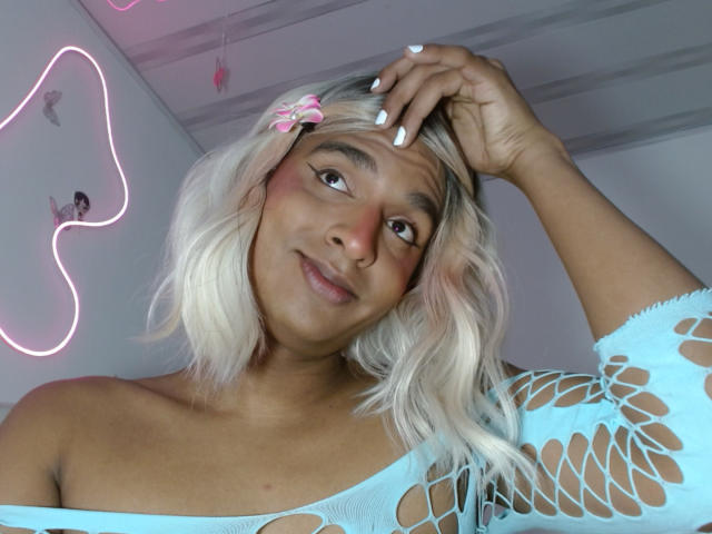 Close-up of IdiiaLatina with blonde hair, wearing a white fishnet top, hand on head, smiling, neon pink outline on white wall, slight blush, confident expression, sexy, casual pose.