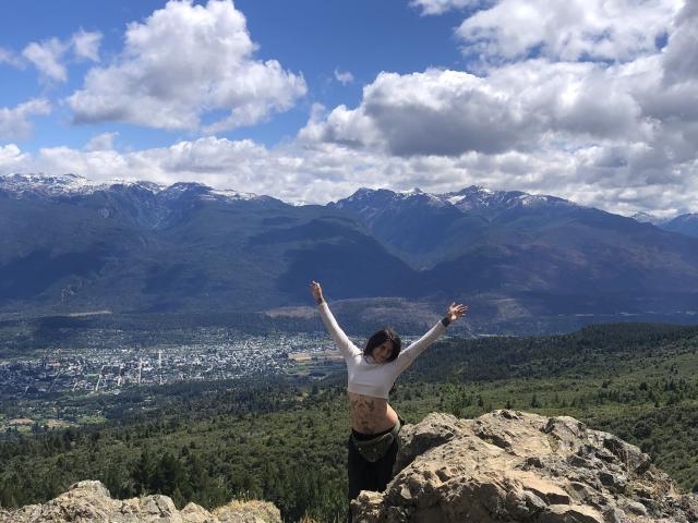 MilaDamian stands on a rocky outcrop with arms raised, facing away. She wears a white long-sleeve top, black pants, and a tattoo on her lower back. Snow-capped mountains and a forested valley below. Bright blue sky with fluffy clouds. Fucking beautiful view.