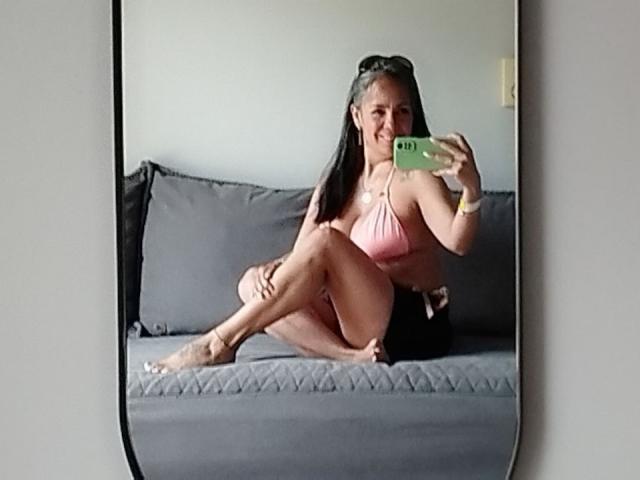 Laila Castillo, ronde, avec de longs cheveux noirs, portant un bikini rose, assise sur un canapé gris, les jambes croisées, tenant une un téléphone vert pour un selfie devant un miroir. Tension sexuelle, attitude confiante, pose détendue..