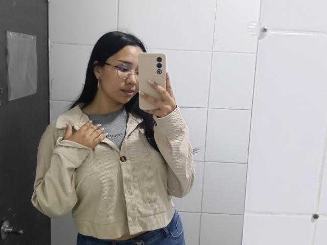 Karollteyes steht vor einer weißen, mit Fliesen verkleideten Wand und macht ein Selfie mit einem rosa iPhone. Sie trägt eine Brille, einen beigen Pullover, ein graues Oberteil und blaue Jeans und lächelt. Total süß..