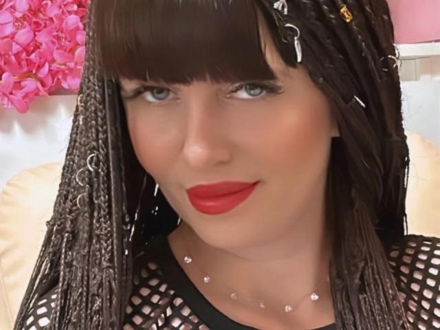 Close-up van CutieOrgasm met bleke huid, zwarte dreadlocks, rode lipstick en een zwarte kanten top, waarbij meerdere piercings in de nek zichtbaar zijn. met felroze bloemen op de achtergrond, kijkend recht in de camera.