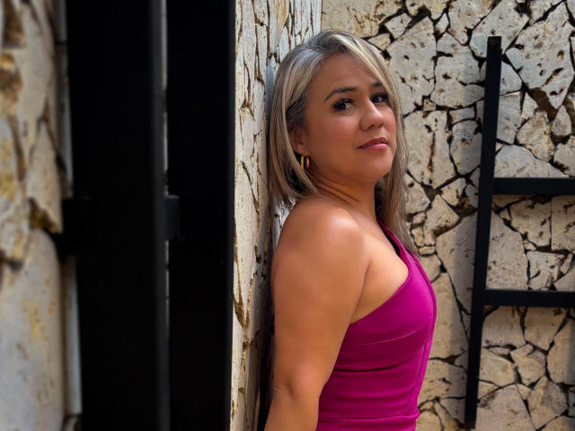 Fotografia de CamilaNoer com cabelo loiro na altura dos ombros, vestindo uma regata rosa justa, encostada em uma parede rachada. Parede de pedra.
ela tem um olhar sedutor, com um leve sorriso. O fundo inclui uma moldura de metal preta..