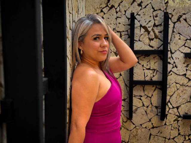 Photographie de CamilaNoer avec des cheveux blonds mi-longs, portant un débardeur rose moulant, appuyée contre un mur fissuré. mur de pierre.
elle regarde l'appareil photo avec un léger sourire, une main dans les cheveux..