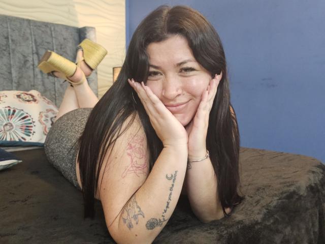 Ayleen Miller, deitada de bruços em uma cama preta, com as pernas levantadas, vestindo shorts brilhantes e saltos altos dourados, sorrindo com as mãos emoldurando seu rosto. Tatuagens visíveis nos braços. Incrivelmente sexy..