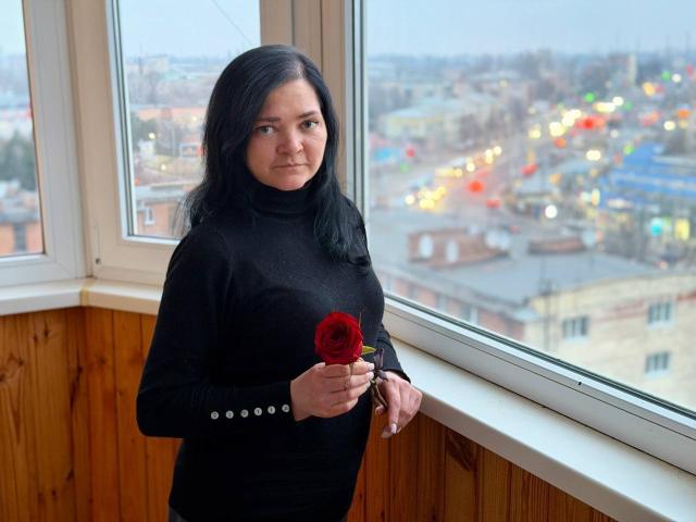 LauraFinn with dark hair, wearing a black turtleneck, holding a red rose in her right hand, standing by a window, wooden panel walls, city lights in the background.