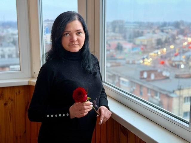 LauraFinn with dark hair, black turtleneck, holding a red rose in her right hand, standing by a window, looking slightly to the left, wooden wall in background.