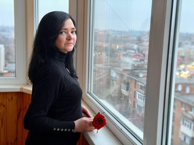 LauraFinn with black hair, wearing a black turtleneck, holding a red rose, standing by a window, looking outside, her tits slightly visible under the tight top.