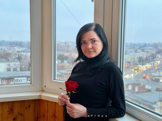 LauraFinn with black hair, holding a red rose, wearing a black turtleneck, standing by a window, wooden paneling, city view, overcast sky, medium breast size, subtle smile.
