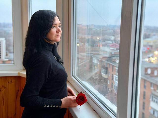 LauraFinn with black hair, wearing a black turtleneck, holding a red rose, standing by a window, looking outside, wooden wall on left, overcast day, cityscape visible.