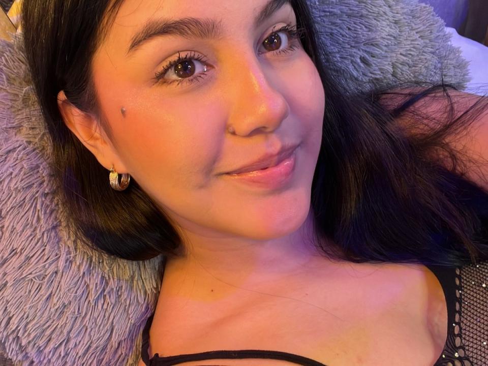 Close-up selfie of AnnyReeseX, lying down with long black hair, brown eyes, and a subtle smile. She wears a black lace top, and a gray textured blanket is in the background.