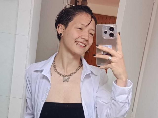 LissyLuxe maakt een selfie met een zilveren iPhone en glimlacht. Ze heeft kort zwart haar, zwarte oorbelgaten, een wit, open shirt, een zwarte top en een ketting. Heel schattig.