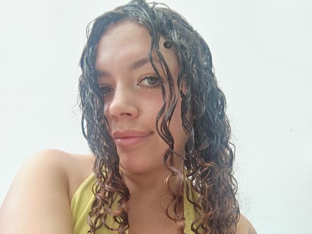 Primo piano di RubbyStrongX, con capelli ricci e neri bagnati, pelle color nocciola, canotta gialla, labbra leggermente dischiuse, sguardo seducente. un leggero sorriso, la luce del sole illumina il viso, tensione sessuale, atmosfera di un selfie casual..