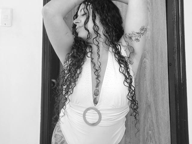 Zwart-wit foto van RubbyStrongX met lang, krullend zwart haar, armen omhoog, gekleed in een witte jurk met een lage hals. Ronde riem. Blootgestelde rug, intense blik, sterke seksuele spanning.