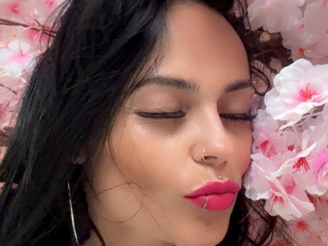 Fotografía de primer plano de CharlotteCrofort con cabello negro, ojos cerrados y lápiz labial rojo, rodeada de flores de cerezo rosadas. Ella tiene un piercing en la nariz y grandes aros, irradiando una sensual tensión..