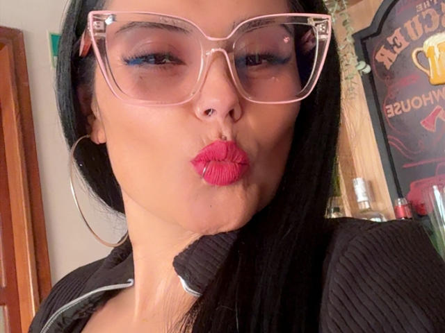 Gros plan de CharlotteCrofort, portant de grandes lunettes transparentes, des cheveux noirs et un rouge à lèvres rouge vif, qui fronce les lèvres. Elle porte un haut noir côtelé, avec un fond uni. Incroyablement sexy..