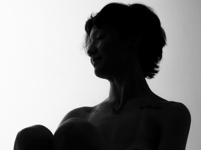 Silhouette de LissyLuxe, une femme avec des cheveux courts et en désordre, de grandes seins, vue de profil, les yeux fermés, le cou orné. avec un collier délicat. Fort contraste, couleurs sombres sur fond clair, forte tension sexuelle..