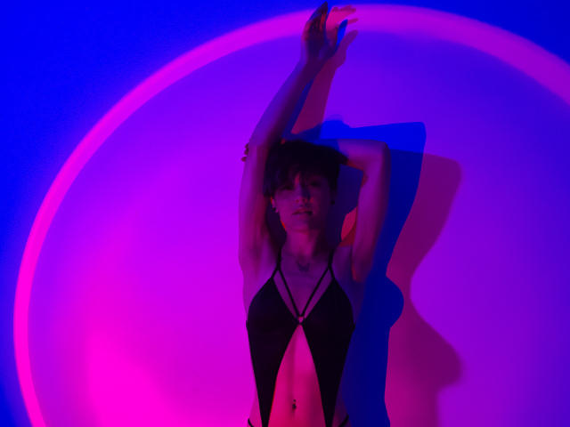 LissyLuxe, mit erhobenen Armen, steht vor einem leuchtenden, rosa und blauen Kreis. Sie trägt ein schwarzes, trägerloses Top, das ihren Bauch freilegt. Neonlichter erzeugen eine verführerische, clubähnliche Atmosphäre..