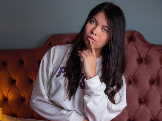 Photographie de CamiiVasquez, avec de longs cheveux noirs, portant un sweat-shirt blanc, assise sur un canapé orange en velours capitonné. Elle se mordille le doigt, et regarde directement la caméra avec une expression séduisante..