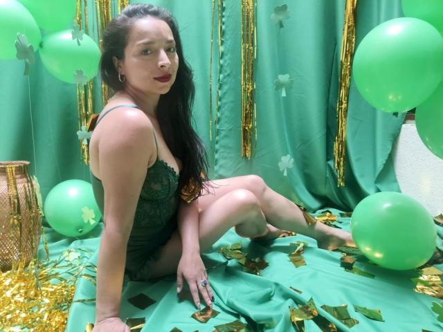 StangelarRivera está sentada sobre una tela verde, rodeada de globos verdes y confeti dorado. Ella lleva una blusa negra de encaje, cabello negro largo y lápiz labial rojo. Su pene no es visible. Pose seductora y juguetona..