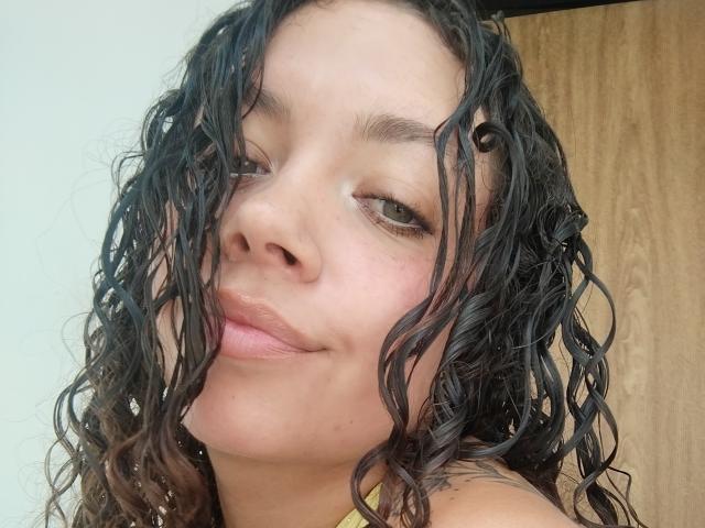Foto em close-up de RubbyStrongX, uma mulher com cabelo preto encaracolado, pele marrom clara e lábios cheios. Ela está olhando ligeiramente para o lado, com um sorriso discreto, e a luz natural ilumina seu rosto..
