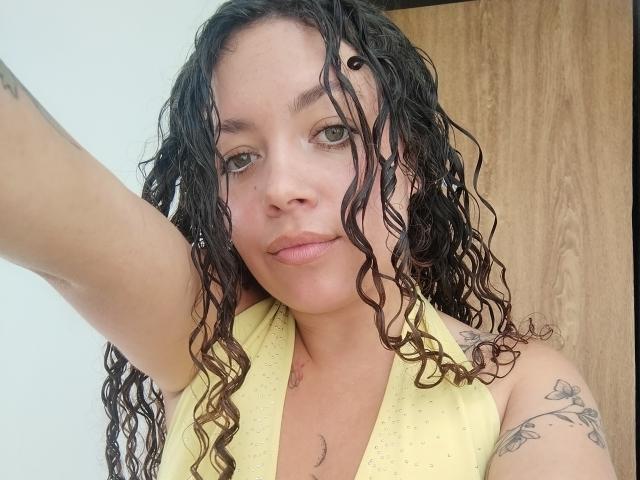 Primo piano di RubbyStrongX, una donna con i capelli bagnati, la pelle chiara e tatuaggi, che indossa un top giallo a tracolla. Sta alzando il braccio destro, guardando direttamente la telecamera con un sorriso appena accennato..