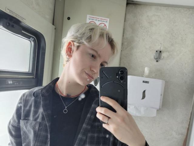 Dionne Szczygiel mit kurzen, blonden Haaren macht ein Selfie in einer öffentlichen Toilette, hält ein schwarzes Telefon und trägt ein schwarzes Karomuster. Jacke, und lächelte leicht. Hand, die ein Telefon hält, weißer Handtrockner im Hintergrund..