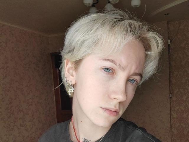 Fotografia de Dionne Szczygiel com cabelo loiro curto e desalinhado, olhos azuis e uma expressão séria. Vestindo uma camisa preta, um colar vermelho e brincos dourados. Quarto com pouca luz e papel de parede com motivos florais..