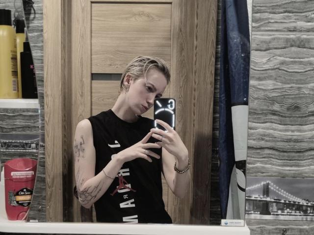 Dionne Szczygiel, con i capelli biondi umidi, tatuaggi sul braccio sinistro, maglietta nera senza maniche, che tiene un telefono, selfie allo specchio, sfondo in legno. sfondo del pannello, bottiglia rossa sul bancone, skyline riflesso nello specchio.