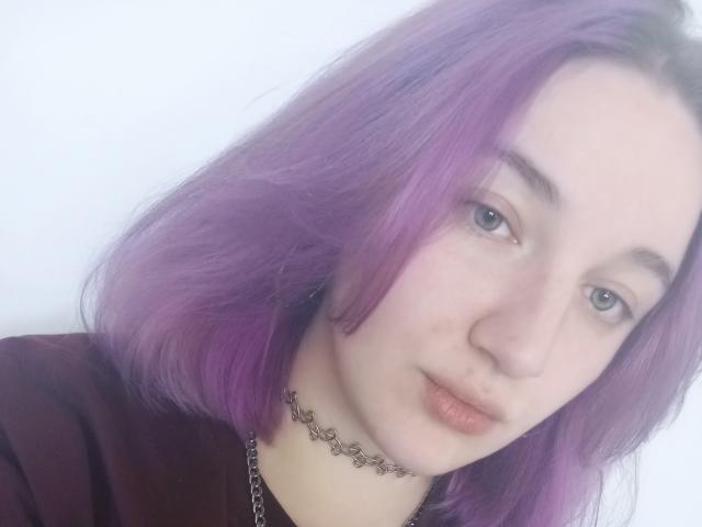 Primo piano di AnnitaMilliter con la pelle chiara e i capelli viola, che indossa una maglietta nera e una collana argentata. Lei guarda direttamente la fotocamera con un'espressione leggermente neutra, e ha gli occhi azzurri. Molto attraente..