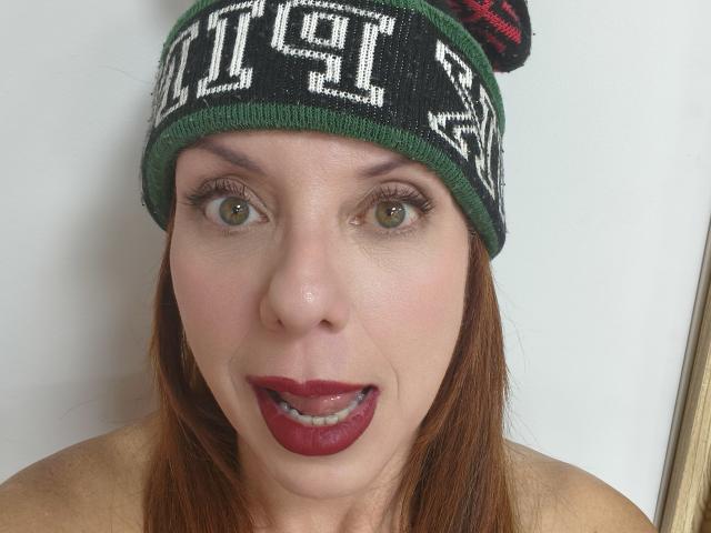 Close-up de Khharha com olhos verdes grandes, batom vermelho e um gorro verde com texto branco. Seu rosto está levemente brilhante, ela está sem blusa e tem uma expressão sexy e excitada..