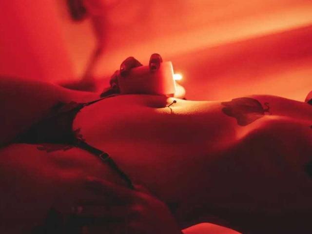 Le torse nu de DakotaRousse, éclairé par une lumière rouge intense, révèle ses grandes seins et une main agrippant un pénis. des ombres soulignant ses courbes et créant une atmosphère sensuelle et érotique..