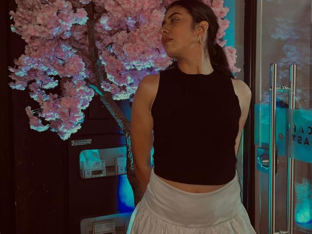 MiluSky mit dunklem Haar, schwarzem Top ohne Ärmel, weißer Rock, steht vor einem leuchtend rosa Kirschbaum, beleuchtet mit blauem Licht. Hintergrund. Hände hinter dem Rücken, leichte Drehung, verführerischer Ausdruck. Verdammt sexy..