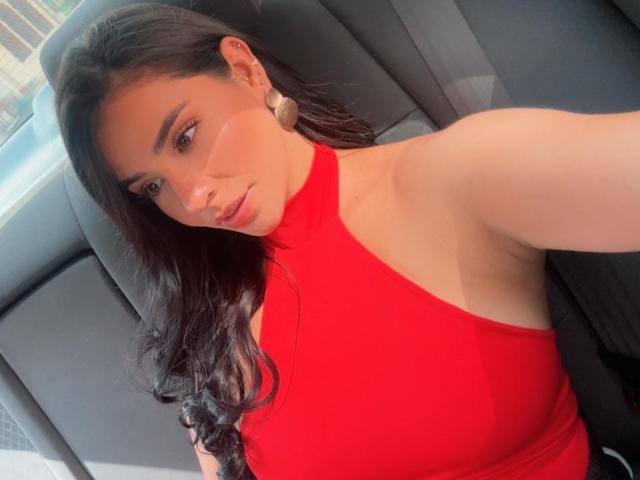 AmaiaCooper, una chica atractiva con el pelo largo y negro, se toma un selfie en un coche mientras lleva una camiseta ajustada de color rojo brillante y sin mangas. Ella tiene grandes aros y una mirada seductora..