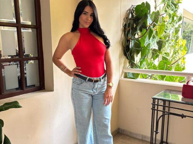 AmaiaCooper, con su largo cabello negro, se encuentra con confianza en una habitación iluminada por el sol, vistiendo un top rojo de tirantes y jeans azules. Tiene una figura atractiva y curvilínea, y una sonrisa arrogante. Hay una planta en maceta en el fondo..