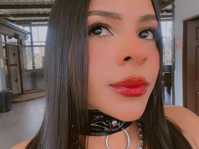 Nahaufnahme von LindaFranco mit dunklem, glattem Haar, rotem Lippenstift und einer schwarzen Lederkette mit silbernen Ringen. Ihre großen, braunen Augen sind intensiv nach vorne gerichtet. Industrieller Hintergrund mit Metallträgern..