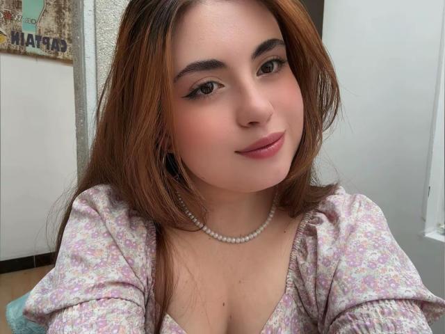 VicluX has fair skin and long brown hair, wearing a floral top with a pearl necklace. She has a slight smile, looking directly at the camera, with subtle makeup. Fucking sexy.