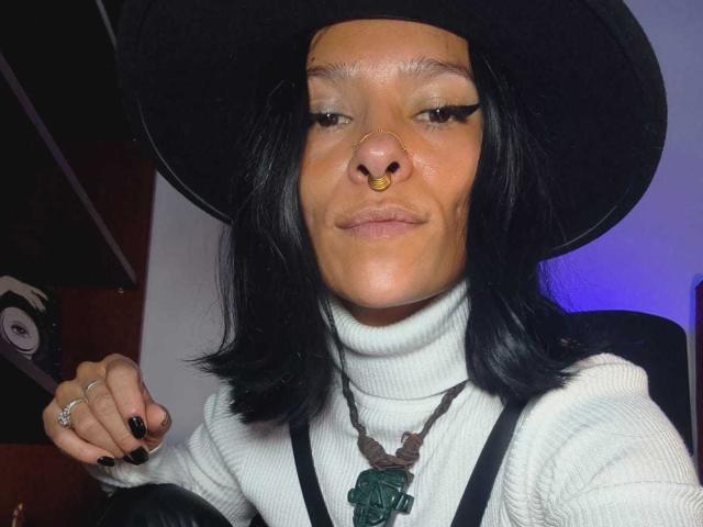 Close-up of AmelieBlake with black hair, wearing a black hat, white turtleneck, black suspenders, and a green pendant. Gold nose ring, black nails, smug expression, blue lighting in background.
