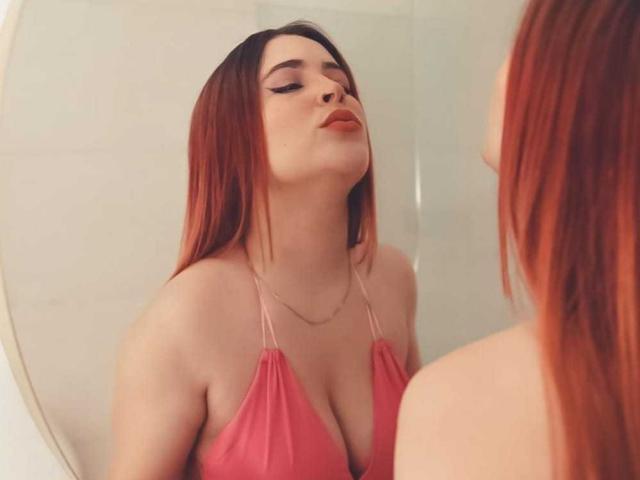Kendra Gill, une femme à la peau claire avec de longs cheveux roux, observe son reflet dans un miroir. Elle porte un haut rose à encolure plongeante, qui révèle une poitrine généreuse. Elle suce ses lèvres, avec un regard séduisant..