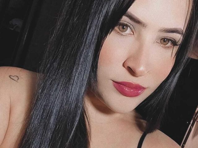 Fotografia ravvicinata di KendraGill con lunghi capelli neri, pelle chiara e rossetto rosso. Lei guarda direttamente la fotocamera con un leggero sorriso. Piccolo tatuaggio a forma di cuore sulla spalla sinistra. Molto attraente..