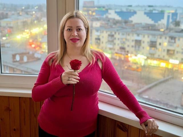 OliviaVerr, eine blonde Frau mit heller Haut, trägt einen roten Pullover mit Reißverschluss und hält eine rote Rose, während sie sich an ein Fenster lehnt. Der Hintergrund zeigt eine Stadtansicht mit verschwommenen Lichtern. Verdammt sexy..